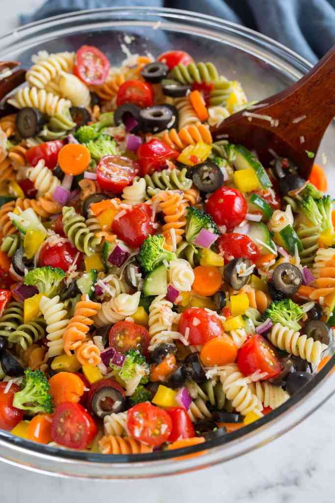 Colorful noodles in a glass bowl with tomatoes, onions, olives, broccoli and dressing. A wooden spoon is scooping it.